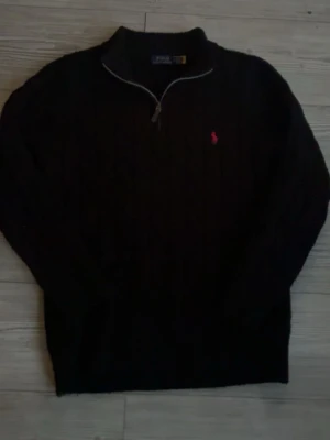 Svart stickad tröja Polo Ralph Lauren - Svart stickad tröja från Polo Ralph Lauren med halv dragkedja och klassisk kabelstickning. Tröjan har en röd broderad logga på bröstet och ribbade muddar vid ärmslut och nederkant. Perfekt för en stilren och avslappnad look.