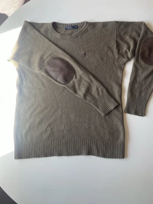 Olivgrön stickad tröja Polo Ralph Lauren - Snygg olivgrön stickad tröja från Polo Ralph Lauren med rund halsringning och bruna mockadetaljer på armbågarna. Tröjan har ribbstickade muddar och en liten broderad logga på bröstet. Perfekt för en stilren och avslappnad look. Samt att tröjan har krympt 2 storlekar. Originellt i storlek M men passar Xs - S beroende på långd, allt över 175cm är tröjan för liten på, så super bra till den som inte är så lång