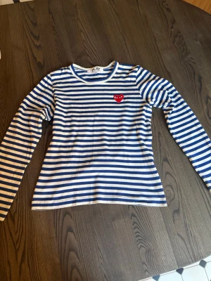 Randig långärmad topp Comme des Garçons - Säljer en blåvit randig långärmad topp från Comme des Garçons Play med det ikoniska röda hjärtat broderat på bröstet. Rund halsringning. Perfekt inför sommaren