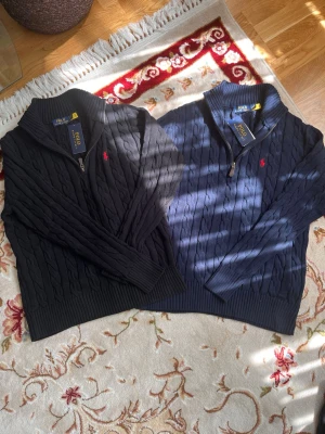 Polo Ralph Lauren Cable-Knit Quarter-Zip (Bundle x2) - Kabelstickad mörkblå tröja från Polo Ralph Lauren med halv dragkedja och röd broderad logga på bröstet. Tröjan har hög krage och ribbstickade muddar. Perfekt för en stilren och klassisk look.