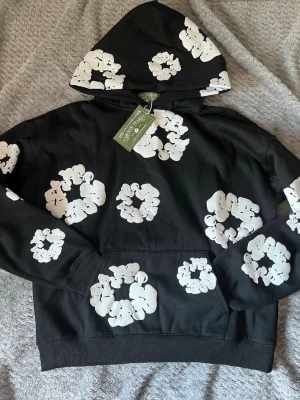 Svart hoodie med vita blommönster - Säljer en svart hoodie från Market med stora vita blommönster tryckta över hela plagget. Hoodien har en klassisk huva och magficka framtill. Materialet känns mjukt och bekvämt, perfekt för en avslappnad streetwear-look.