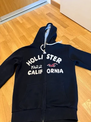 Marinblå Hollister hoodie med dragkedja - Mörkblå hoodie från Hollister med vit broderad text och logga på bröstet. Dragkedja framtill, två fickor och vit dragsko i huvan. Insidan av huvan är ljusgrå. Perfekt för en avslappnad stil. 
