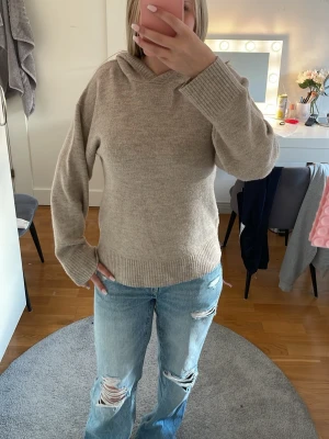 Beige stickad hoodie - Mysig beige stickad hoodie med avslappnad passform och ribbade muddar. Tröjan har huva och är perfekt för lager-på-lager. Passar dig som gillar en enkel och stilren look. Materialet ser ut att vara mjukt och skönt, perfekt för kyligare dagar.