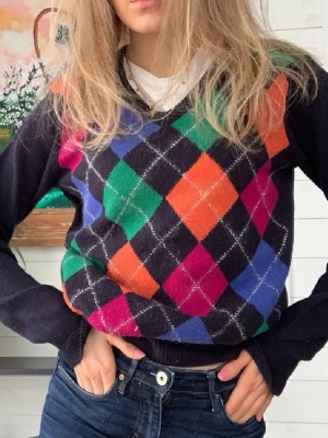 Färgglad argyle stickad tröja - Stickad tröja med klassiskt argylemönster i grönt, blått, orange och cerise på svart botten. Tröjan har v-ringning och långa ärmar i svart. Perfekt för dig som vill sticka ut med färg och mönster. Materialet ser ut att vara mjukt och mysigt.