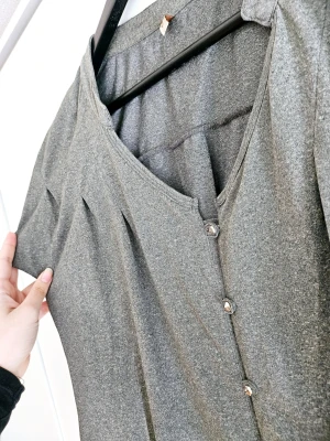 Grå vekad blus med knappar - Snygg grå vekad blus med v-ringning och korta ärmar. Blusen har dekorativa knappar framtill och ett lätt, luftigt fall. Perfekt för dig som vill sticka ut lite extra med en touch.  