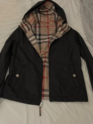 Svart Burberry vindjacka med huva - Stilren svart vindjacka från Burberry med klassiskt rutigt foder i beige, rött och svart. Jackan har huva, två fickor med tryckknappar och dragkedja framtill. Märkespatch på bröstet. Perfekt för en trendig och sportig look.