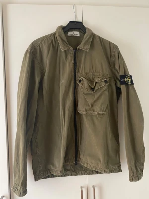 Olivgrön overshirt från Stone Island - Snygg olivgrön overshirt från Stone Island med klassisk patch på ärmen. Jackan har dragkedja framtill, en stor bröstficka med lock och knapp samt krage. Tillverkad i lätt bomullsmaterial, perfekt för lager-på-lager-stil. Riktig snygg inför sommaren!