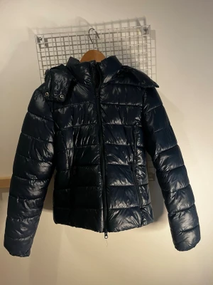 Mörkblå pufferjacka med huva - Snygg mörkblå pufferjacka med glansig finish och avtagbar huva. Jackan har dragkedja framtill, två sidofickor med dragkedja och quiltad design.💕💕