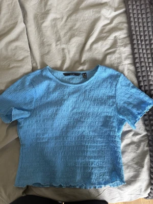 Topp  - En ljusblå topp från vero lofa med korta ärmar och rund halsringning. Toppen har en skrynklig struktur och vågig kant nertill för en trendig look. Perfekt att styla med jeans eller kjol.