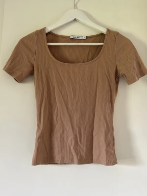 Ribbad beige t-shirt från NA-KD - Säljer en ribbad beige topp från NA-KD med korta ärmar och fyrkantig ringning. Toppen har en slim passform och är tillverkad i mjuk bomullsmix, perfekt för en enkel och stilren look.