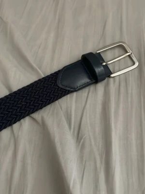Mörkblått flätat bälte med skinn - Snyggt mörkblått flätat bälte med detaljer i skinn och silverfärgat spänne. Perfekt accessoar för att lyfta din outfit med en stilren touch. Bältet har en klassisk design och passar till många olika looks.