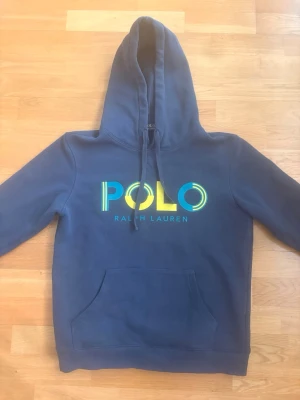 Blå hoodie från Polo Ralph Lauren - Snygg blå hoodie från Polo Ralph Lauren med stor färgglad logga på bröstet. Klassisk modell med huva och dragsko samt en stor magficka. Perfekt för en chill och avslappnad stil. Materialet känns mjukt och bekvämt mot huden. Storleken är M men den sitter som S.