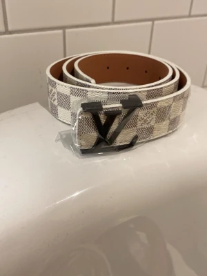 Vit bälte - Snyggt vit bälte med rutigt mönster med en svart logga. Passar till alla clean outfits!