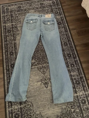 Ljusblå bootcut jeans från True Religion - Säljer ett par ljusblå bootcut jeans från True Religion med klassiska kontrastsömmar och fickor med lock och knappar bak. Jeansen har låg midja och är tillverkade i denim med lätt stretch. Dem är uppsyda längst ner för hand men de går såklart att ta bort,passar perfekt i längden till mig som är 168