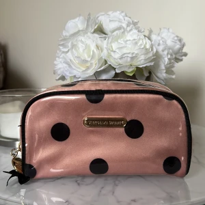 Rosa necessär från Victoria's Secret - Säljer en söt rosa necessär från Victoria's Secret med stora svarta prickar. Väskan har en dragkedja med en guldfärgad detalj och en svart rosett. Perfekt för att hålla ordning på dina småsaker.