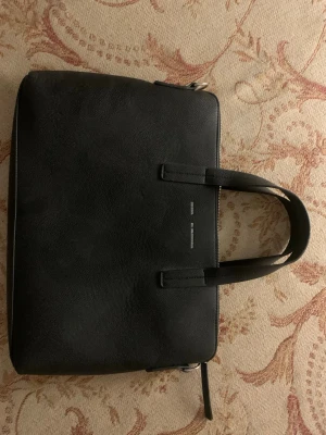Svart datorväska från glitter  - Stilren svart datorväska i slätt läder. Väskan har två handtag, dragkedja upptill och ett rymligt innerfack med extra ficka för småprylar. Perfekt för laptop och viktiga papper. Enkel och klassisk design som passar till allt.