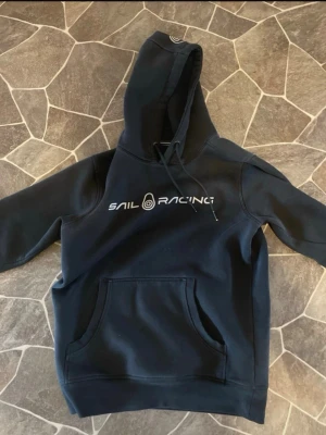 Marinblå Sail Racing hoodie - Snygg marinblå hoodie från Sail Racing med vit logga på bröstet. Klassisk känguruficka framtill och justerbar huva med dragsko. Perfekt för en avslappnad och sportig stil.