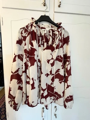 Blommig blus med volangkrage - Snygg blus med vinrött och vitt blommönster, volangkrage och knytband vid halsen. Luftig passform och långa ärmar med resår vid handlederna. Perfekt för dig som gillar en trendig och avslappnad stil.