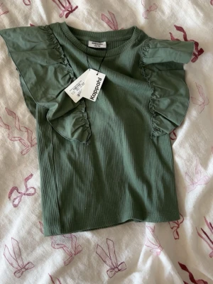 Grön ribbad topp med volangärm Kappahl - Supersnygg grön ribbad topp från Kappahl med volangdetaljer på axlarna. Toppen har rund halsringning och är tillverkad i mjuk bomullsmix. Perfekt för dig som vill ha något extra till din outfit!