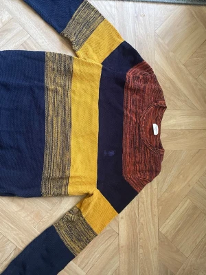Randig stickad tröja från US Polo Assn - Stickad tröja med breda ränder i marinblått, senapsgult och rostrött från US Polo Assn. Tröjan har rund halsringning och broderad logga på bröstet. Perfekt för dig som gillar färg och vill ha en unik look.