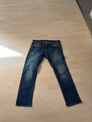 Blå jeans från Replay, W34 L32 - Snygga blå jeans från Replay med klassisk femficksmodell och raka ben. Jeansen har en lätt tvättad look med slitningar på låren och knäna. Perfekta för en avslappnad och trendig stil.