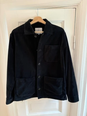 Svart overshirt från Bläck i manchester - Stilren svart overshirt från Bläck i mjuk manchester. Klassisk krage, tre stora fickor framtill och knäppning med knappar. Perfekt lager-på-lager-plagg med avslappnad passform och tidlös look.