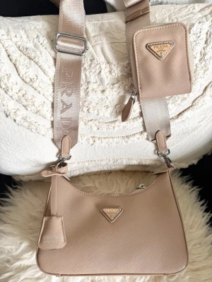 Supersnygg Beige Axelväska - Sällsynt och snygg beige axelväska med silverdetaljer och avtagbar axelrem. Väskan har ett ikoniskt trekantigt pradalogo framtill och en liten matchande miniväska på remmen. Perfekt för dig som vill sticka ut med en ikonisk och lyxig accessoar. Bara använd några gånger, väskan är fortfarande i väldigt fint skick.