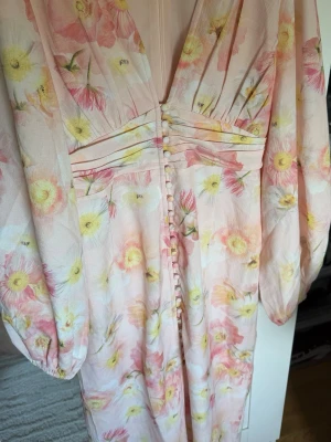 Blommig ballong sleeve - Ballong Sleeve långklänning med pastellfärgade blommor i rosa och gult på ljus botten. Djup v-ringning, långa puffärmar och dekorativa knappar framtill. Markerad midja med veckade detaljer. Perfekt för sommarens tillfällen. Endast testad. 