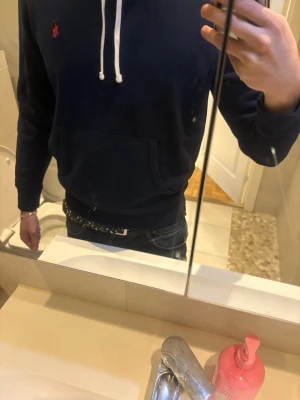 Mörkblå hoodie från Ralph Lauren - Mörkblå hoodie från Ralph Lauren med vit dragsko, känguruficka och röd broderad logga på bröstet. Klassisk passform med ribbade muddar och mjukt material som känns skönt mot huden. Modellen på bilderna är 184 cm / 74 kg för referens på passformen. 