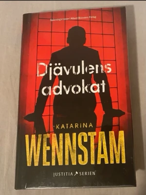 Djävulens advokat av Katarina Wennstam - Spännande kriminalroman i Justitia-serien av Katarina Wennstam. Omslaget är svart och rött med siluett av en person mot ett rutigt fönster och stora gula bokstäver. Inbunden bok på svenska, perfekt för dig som gillar deckare och thrillers med juridiska teman.