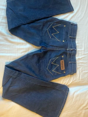 Bootcut lågmidjade mörkblå Wrangler jeans - Säljer ett par mörkblå bootcut jeans från Wrangler med låg midja och klassiska W-sömmar på bakfickorna. Jeansen har gul kontrastsöm och är tillverkade i slitstarkt denim. Perfekt för dig som gillar retrovibbar och vill ha en snygg siluett.