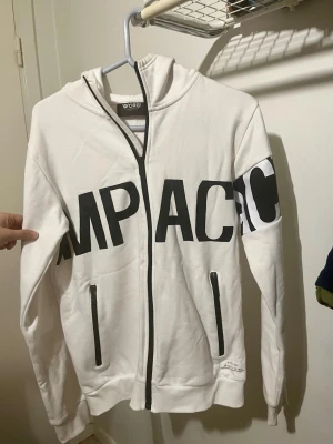 Sweatshirt  - Vit hoodie med dragkedja och texten ”IMPACT” tryckt över bröstet och ärmen från Kappahl i storleken 170. Den har två fickor med dragkedja framtill och luva. 80% bomull. Aldrig använd.