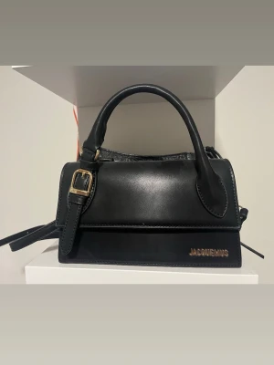 Svart Jacquemus handväska i läder - Snygg svart handväska från Jacquemus i slätt läder med guldfärgade detaljer och ett justerbart axelband. Väskan har ett stilrent och kompakt format med logotypen i guld på framsidan. Perfekt accessoar för att lyfta din outfit. 