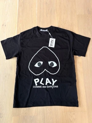 Svart t-shirt Comme des Garçons Play - Svart t-shirt från Comme des Garçons Play med det ikoniska hjärtmotivet och ögon på bröstet. T-shirten har rund hals, korta ärmar och är tillverkad i mjuk bomull. Perfekt för en stilren och trendig look.