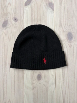 Svart mössa från Polo Ralph Lauren - Svart ribbstickad mössa från Polo Ralph Lauren i mjuk merinoull. Klassisk design med uppvikt kant och röd broderad logga framtill. Perfekt accessoar för kyliga dagar och enkel att matcha med olika outfits.