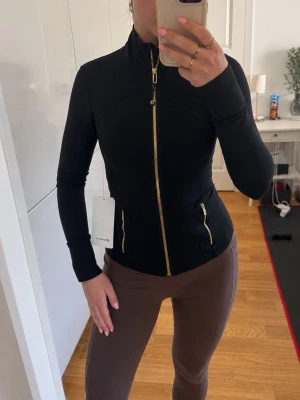 Svart träningsjacka från Lululemon XS define jacket black gold svart guld  - Storlek 2 motsvarar XS , Svart figursydd träningsjacka från Lululemon med guldiga dragkedjor framtill och på fickorna. Jackan har hög krage, långa ärmar och är tillverkad i ett stretchigt funktionsmaterial som passar perfekt till träning eller som lager på lager.