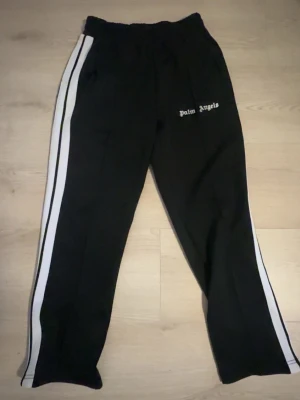 Svarta mjukisbyxor från Palm Angels - Svarta mjukisbyxor från Palm Angels med logga i vitt tryck på benet. Tillverkade i 100% polyester för en skön och sportig känsla. Perfekta för en avslappnad och trendig streetwear-look.