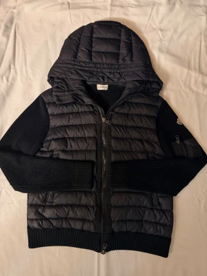  Moncler cardigan  - Snygg svart pufferjacka från Moncler med stickade ärmar och vadderad kropp. Jackan har en stor huva, dragkedja framtill och en liten ficka med dragkedja på ena ärmen. Perfekt för kalla vinterdagar och riktigt trendig look.