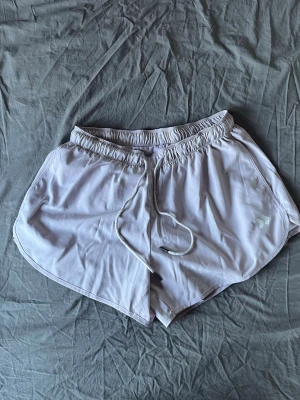 Ljusgrå träningsshorts med snörning - Säljer ett par ljusgrå träningsshorts med elastisk midja och snörning framtill. Shortsen har en sportig look med rundade benslut och är tillverkade i ett lätt syntetmaterial som andas. Har även underbyxor som sitter lite tightare på benen, men med en elastisk och mjuk känsla. Perfekta för gymmet eller löprundan.