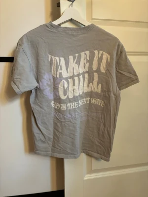 Grå t-shirt med tryck 'Take It Chill' - Grå t-shirt med korta ärmar och rund hals. På ryggen finns ett stort tryck i vitt och lila med texten 'Take It Chill, Catch The Next Wave' och en liten blomma. Materialet är mjuk bomull, perfekt för en avslappnad stil.
