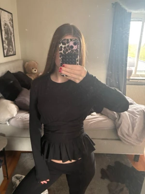 Svart topp med volang från Gina Tricot - Säljer en svart långärmad topp från Gina Tricot med markerad midja och volang nedtill. Toppen har en tight passform och är tillverkad i mjukt material som sitter snyggt på kroppen. Perfekt för dig som vill ha en trendig och stilren look.