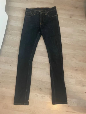 Mörkblå jeans från Nudie Jeans - Säljer ett par mörkblå jeans från Nudie Jeans med klassisk femficksdesign och kontrastsömmar. Jeansen har rak passform och är gjorda i slitstark denim med läderpatch bak i midjan. Perfekta för dig som gillar stilrena och tidlösa jeans. Jeansen är extremt bra skick! Hör av dig om du har några funderingar!🤩