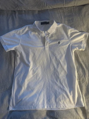 Vit pikétröja från Ralph Lauren - Klassisk vit pikétröja från Ralph Lauren med kort ärm och krage. Tröjan har knappar framtill och den ikoniska broderade loggan på bröstet. Perfekt till jeans eller shorts för en clean och stilren look.