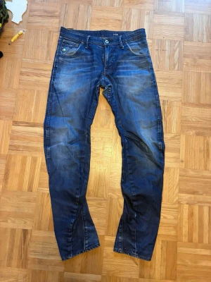 Blå jeans från G-Star med tvättad look - Säljer ett par snygga blå jeans från G-Star med cool tvättad effekt och markerade sömmar. Jeansen har fem fickor, raka ben och klassisk knappgylf. Perfekta för dig som gillar streetstyle och vill ha en avslappnad men trendig look.