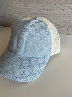 Ljusblå Gucci keps med mesh - Snygg ljusblå keps från Gucci med klassiskt GG-mönster över hela framsidan. Baksidan är i vit mesh för extra ventilation. Perfekt accessoar för en trendig och avslappnad stil. Justerbar passform och böjd skärm.