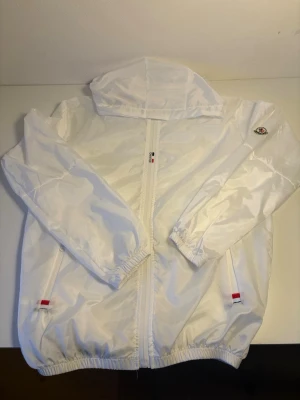 Vit vindjacka  - Snygg vit vindjacka från Moncler med huva och dragkedja framtill. Jackan har elastiska muddar vid ärmar och nederkant samt två fickor med röda och blå detaljer. Moncler-logga på vänster ärm. Perfekt för blåsiga dagar.