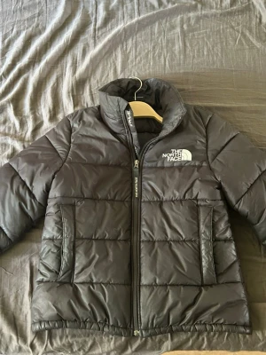 Svart pufferjacka från The North Face - Snygg svart pufferjacka från The North Face med hög krage och dragkedja framtill. Jackan har två stora fickor på sidorna och logga på bröstet. Perfekt för kalla dagar tack vare det vadderade materialet.