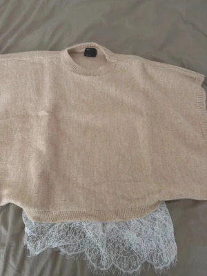 Beige poncho med spetsdetalj ASOS  - Säljer en beige stickad poncho från ASOS med rund halsringning och bred passform. Nedtill finns en snygg vit spetskant som ger en unik och trendig look. Perfekt att slänga över en enkel outfit för att lyfta stilen.