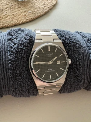 Tissot PRX Powermatic 80 klocka - Snygg Tissot PRX Powermatic 80 klocka med svart urtavla och datumvisning. Nyskick med skyddsplast kvar. Perfekt för dig som vill ha en stilren klassisk klocka utan att tömma plånboken.