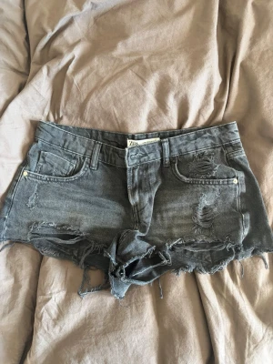 Grå jeansshorts från Zara - Grå jeansshorts från Zara med slitningar. Lågmidjade och perfekta på sommaren.